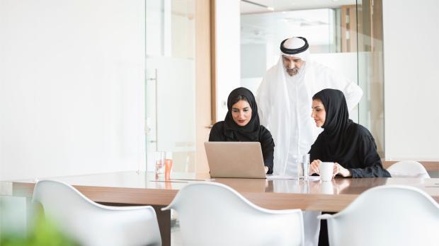 A group of Emirati professionals working together on a laptop in a bright modern office, representing collaboration and tailored employee benefits in the UAE.
