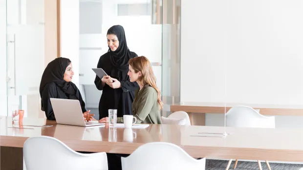 UAE professionals collaborating in a modern office Three female professionals, including Emirati women in abayas, discussing employee benefits in a bright modern office — representing collaboration and engagement in the UAE workplace.